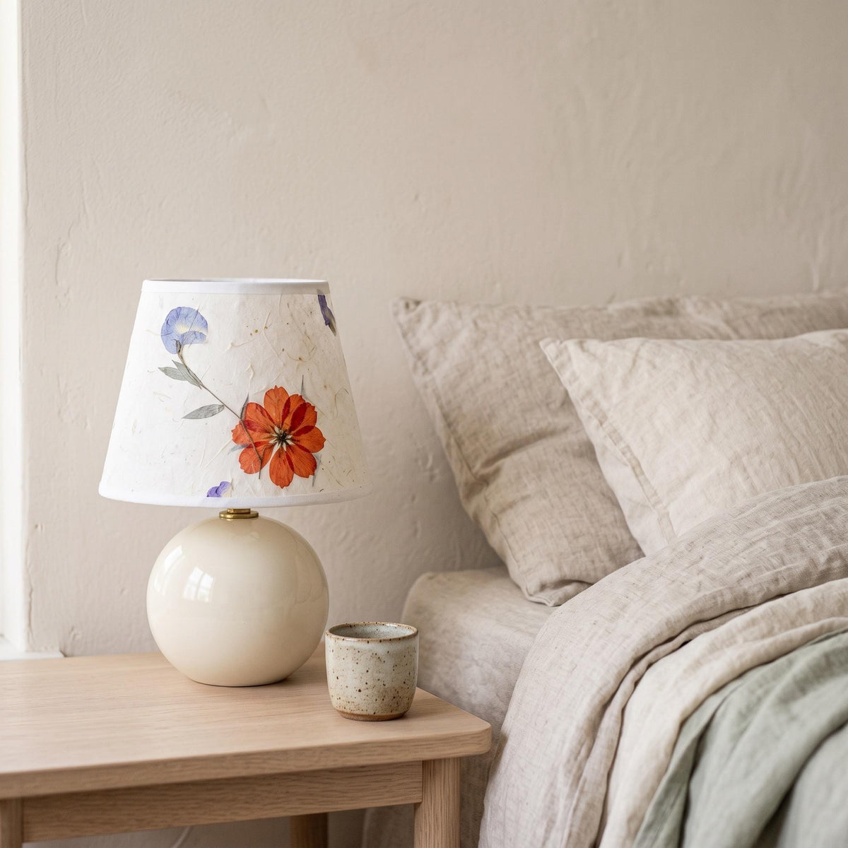 Pressed flower lamp on a bedside table in morning light beside a small cream cup and white linen bedding