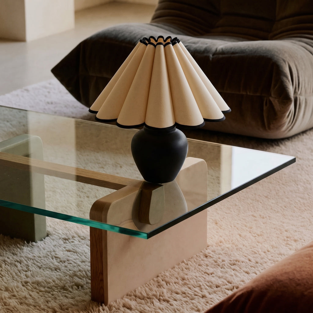Matte black round-bellied lamp base with a cream petal pleated shade piped in black, on a glass coffee table with a wooden block base