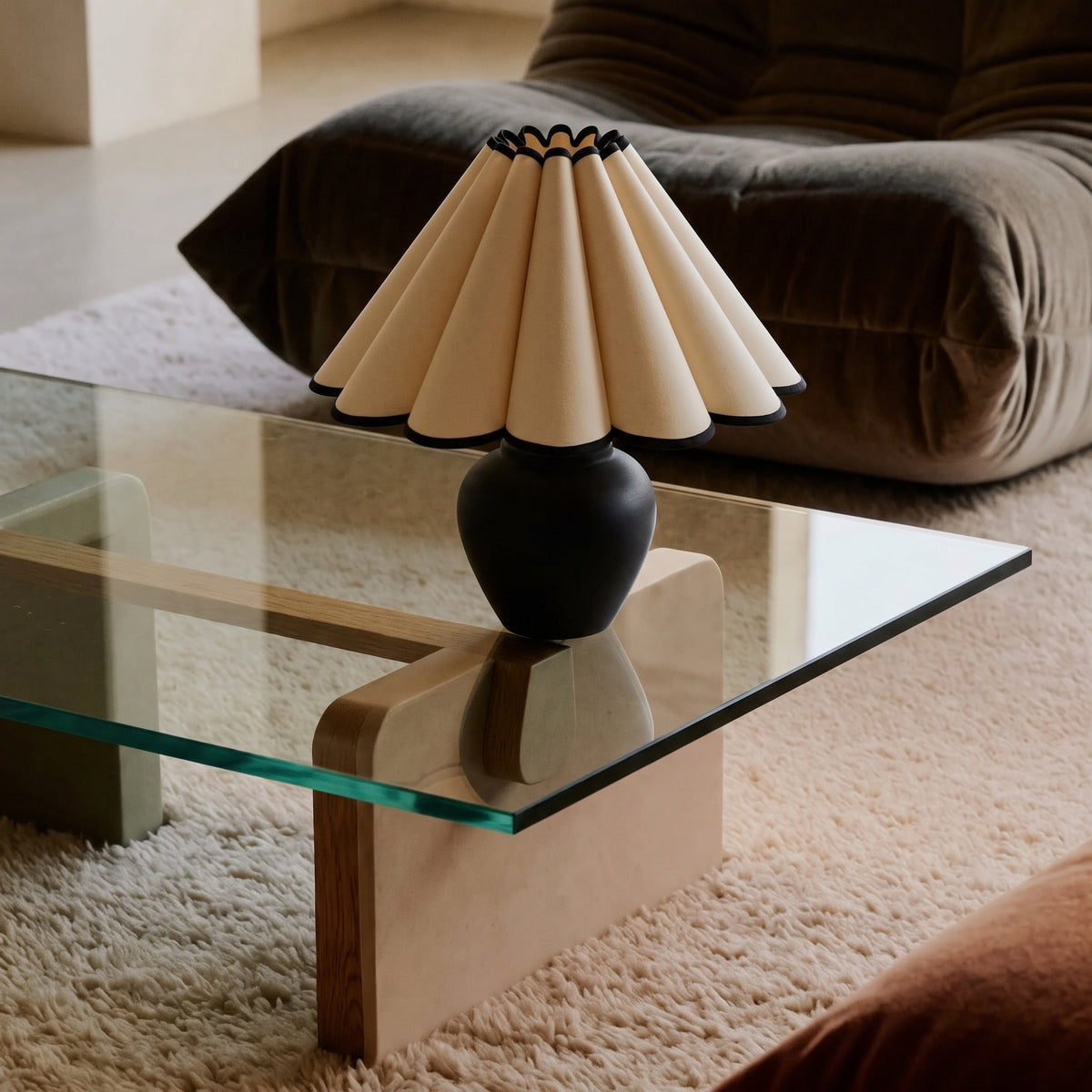 Matte black round-bellied lamp base with a cream petal pleated shade piped in black, on a glass coffee table with a wooden block base