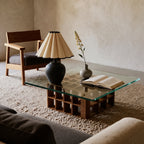 Matte black round-bellied lamp with a cream petal pleated shade on a glass coffee table with a wooden grid base, a small grey vase and an open book beside it