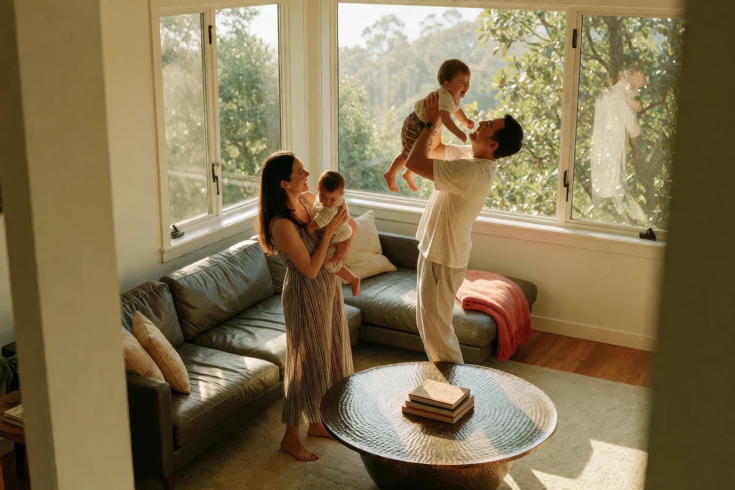 A father lifts a laughing toddler while the mother holds a baby, in a sunlit living room with large windows